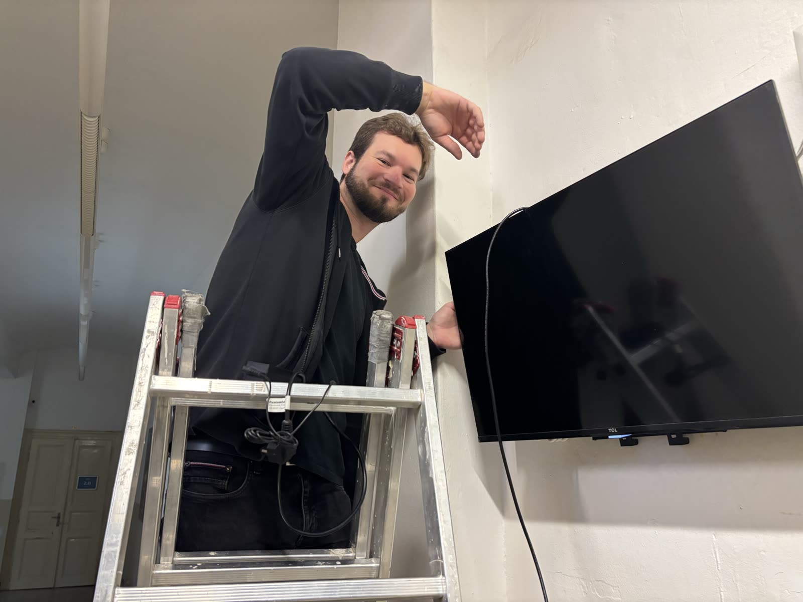 Technician on a ladder hanging the TV on the wall bracket, power cable running from the TV