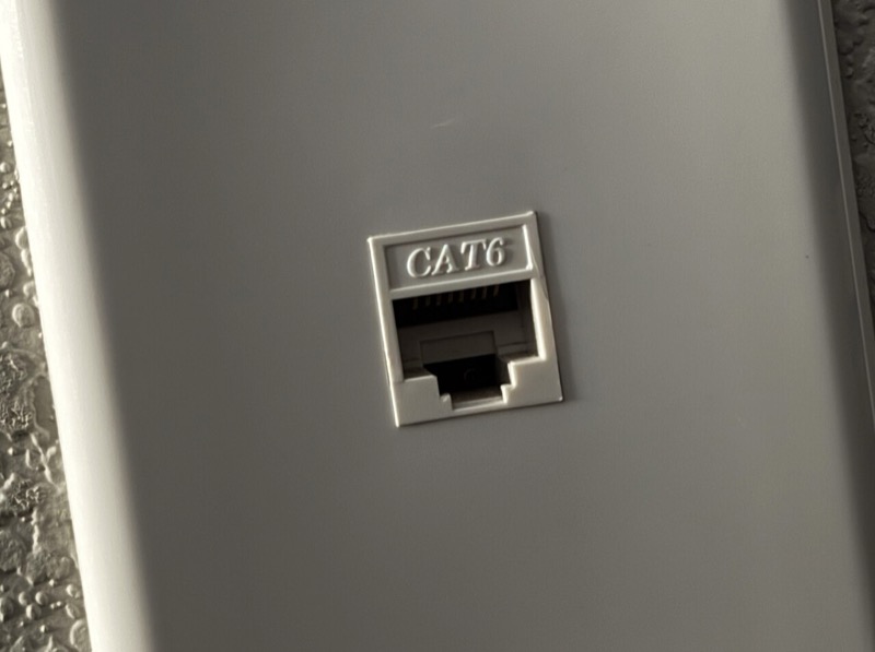 Síťová zásuvka RJ45 (ilustrační foto: Tony Webster, CC BY 2.0, Wikimedia Commons) Bílá síťová zásuvka na zdi s jedním konektorem RJ45 a nápisem CAT6