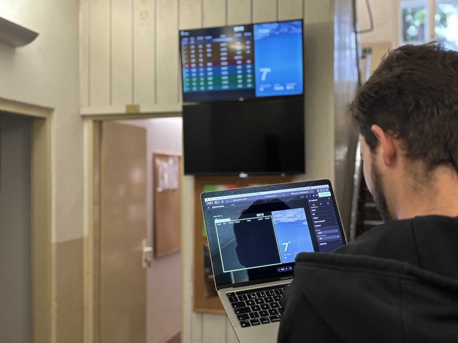 Pohled přes rameno na notebook s otevřeným Studiem, nad dveřmi visí obrazovka se suplováním a počasím
